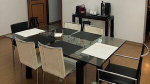 Un comedor con mesa de vidrio, sillas negras, una lámpara de araña y una zona de cocina con electrodomésticos.