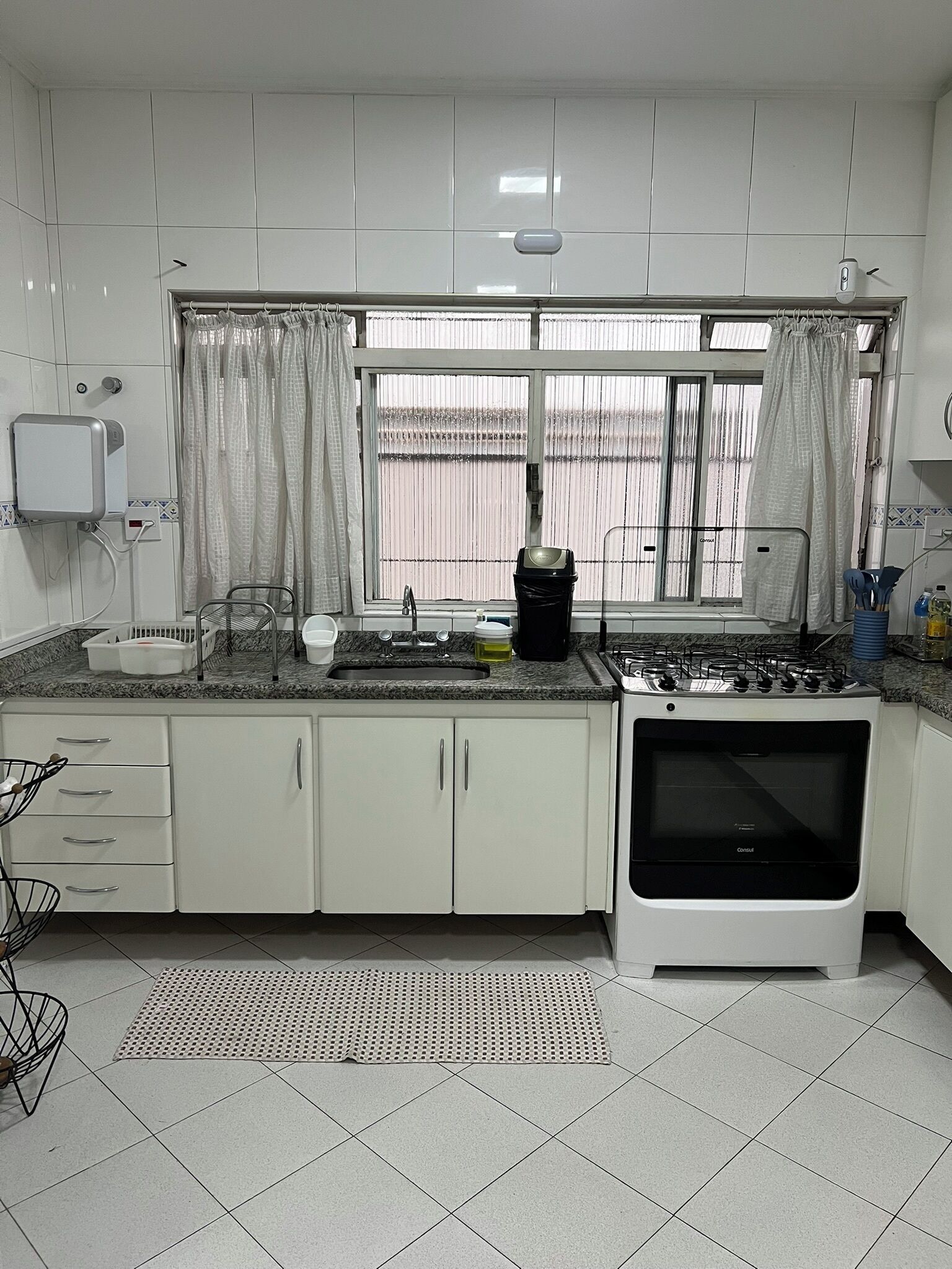 Une cuisine avec des meubles blancs, une cuisinière à gaz, un évier et une fenêtre avec des rideaux.