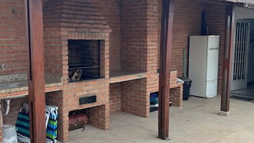 Uma cozinha ao ar livre construída em tijolos, com chaminé, um pátio ladrilhado e uma geladeira branca ao fundo.
