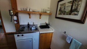 A compact kitchen with a stove, sink, and a small refrigerator. There is a wooden shelf above the sink holding dishes and glasses. A framed picture hangs on the wall to the right. - Cozy stay at Haliburton Forest. Trails, Wolf Centre, Canopy Tour, 100 lakes! (Dysart and Others)