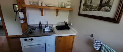 Een praktische keuken met een kookplaat, spoelbak en een kleine koelkast. Daarboven, boven de spoelbak, bevindt zich een houten plank met servies en glazen. Een ingelijst beeld hangt aan de rechterkant van de muur.