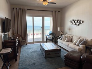 A living room with a sofa, armchairs, a coffee table, a television, and a view of the beach through the sliding glass door.