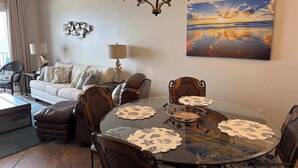 Ein Wohnzimmer mit rundem Glastisch, gepolsterten Sesseln, einer Couch, einer Lampe und einem Gemälde von einem Sonnenuntergang.