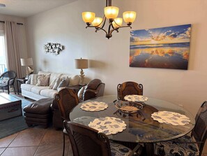 Ein Wohnzimmer mit rundem Glastisch, gepolsterten Sesseln, einer Couch, einer Lampe und einem Gemälde von einem Sonnenuntergang.