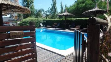 Zona de piscina con terraza de madera, sillas de descanso y sombrillas de paja.