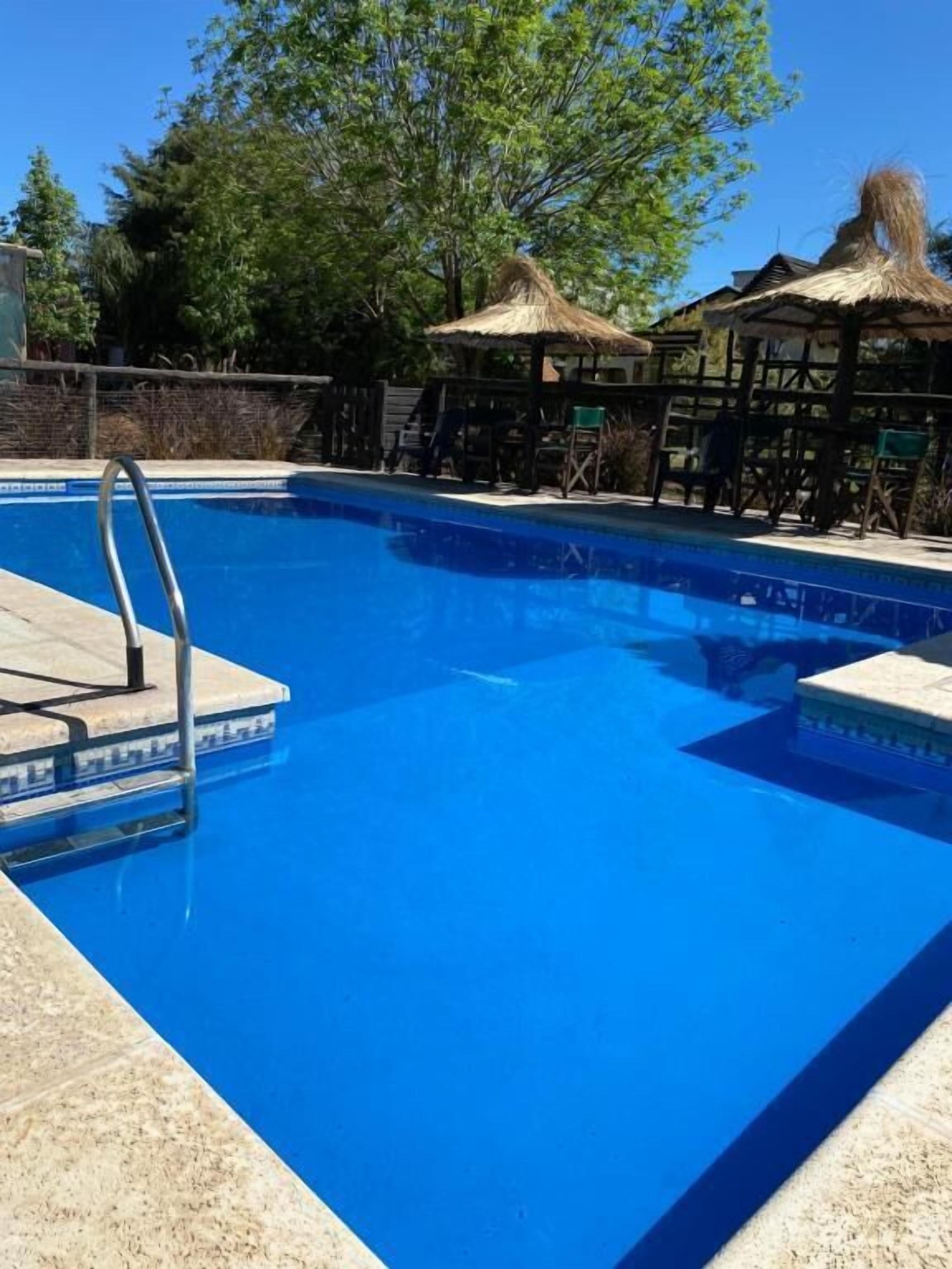 Une piscine avec un escalier, entourée d’une terrasse en bois et de constructions à toits de chaume.