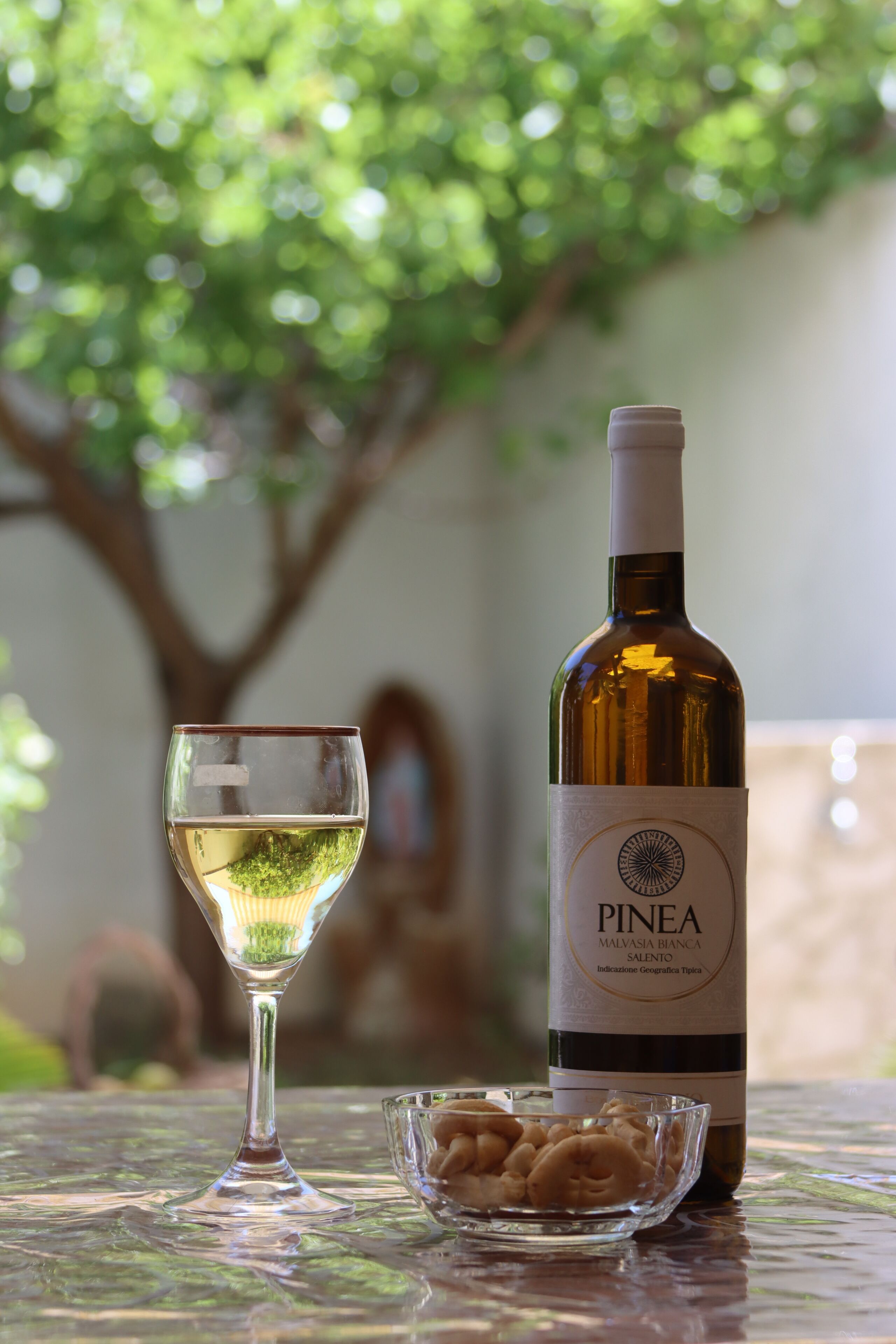 Una botella de vino PINEA y una copa de vino blanco sobre una mesa con un cuenco de vidrio lleno de frutos secos.
