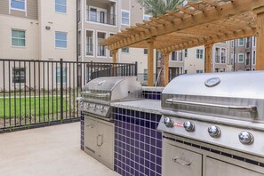 An outdoor grill area with a pergola overhead, a fenced patio, and a view of apartment buildings in the background. - Cozy 1-bedroom apartment in peaceful Houston with AC, pool, and gym access (Houston)