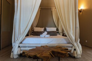 Una cama con dosel y cortinas, ropa de cama blanca y almohadas marrones, una mesa de centro de madera y una lámpara fijada a la pared.