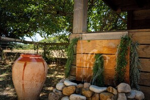 Entorno rústico al aire libre con una estructura de madera, una gran maceta de cerámica y una cascada pequeña rodeada de rocas y follaje.