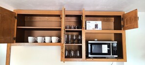 Ein Holzschrank mit offenen Regalen, der weiße Tassen, klare Gläser und eine Mikrowelle beherbergt.