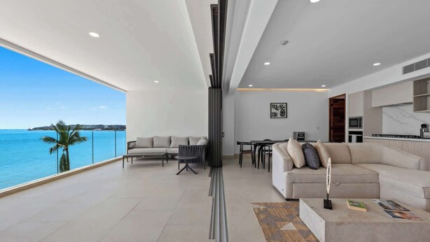 A modern living room with a large window overlooking a body of water, a sofa, a coffee table, and a dining area. - Panarea 2C: Lux Beachfront 3BR, Oceanviews + Pools (Bucerías)
