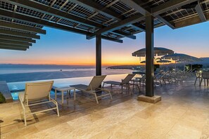 Piscina infinita com espreguiçadeiras e guarda-sóis, com vista para um pôr do sol sobre o oceano.