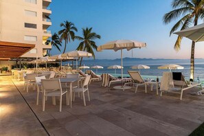 Um terraço à beira-mar com mesas, cadeiras, guarda-sóis e piscina.