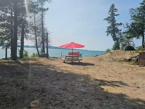 A beach with a red umbrella and a bench, surrounded by trees and a view of the water.
