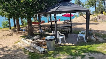 A covered outdoor seating area with tables and chairs, a trash can, and a grill, overlooking a beach with a clear view of the water.