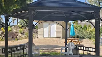 Um gazebo com um banco e cadeiras, uma mesa de piquenique e uma casa ao fundo.