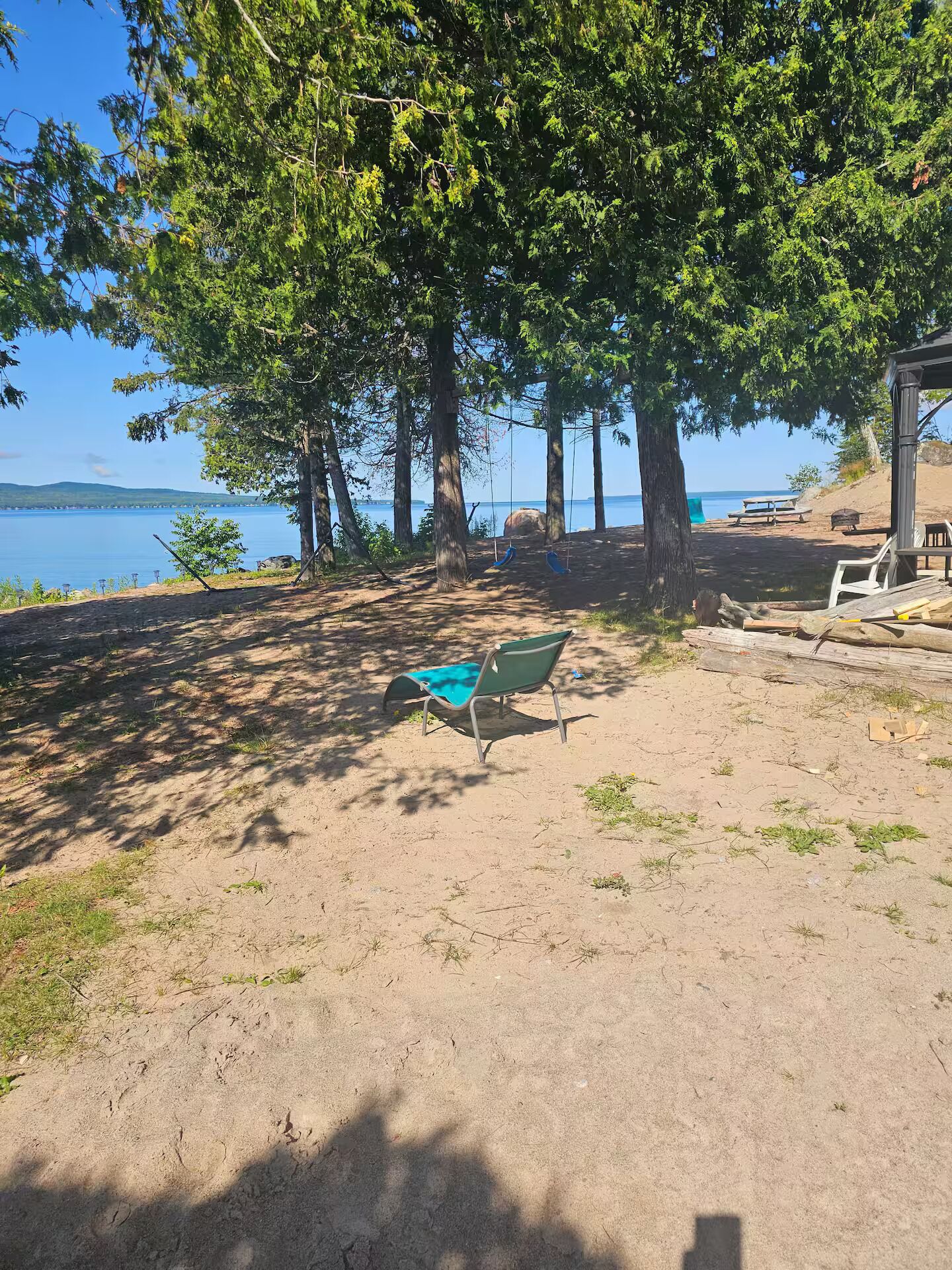 Ein Sandstrand mit einem grünen Stuhl und einer Hängematte unter einem großen Baum, mit Blick auf ein Gewässer.