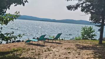 A beach with a row of green lounge chairs facing a calm lake, surrounded by trees.