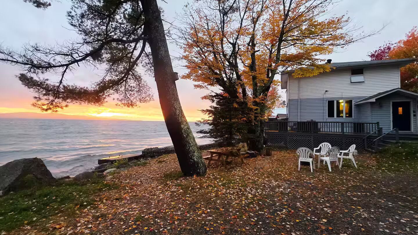 A waterfront property with a house, outdoor seating, and a view of the ocean at sunset.