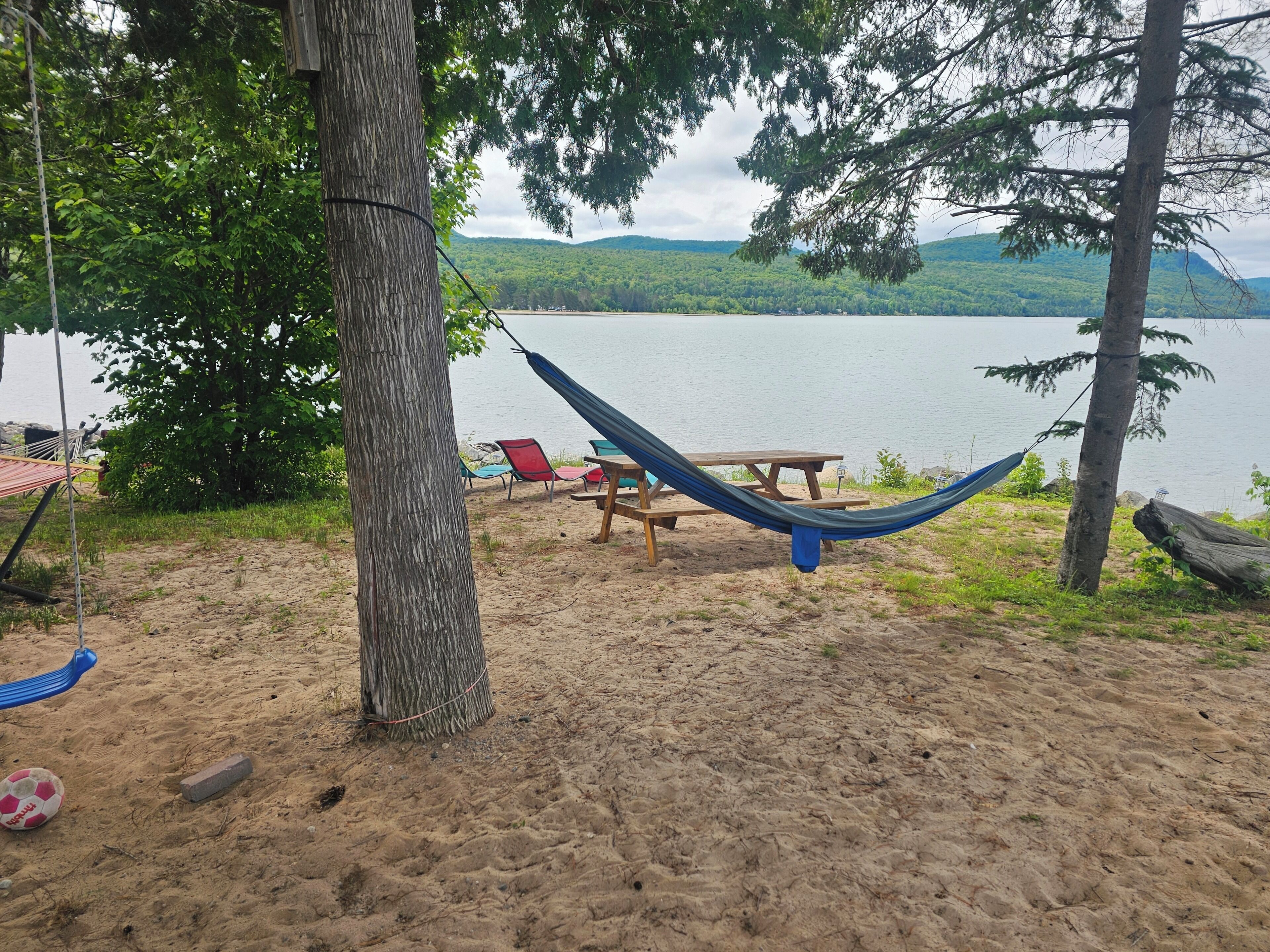 Eine Hängematte, die an einem Baum befestigt ist, ein Picknicktisch und ein roter Stuhl am See.