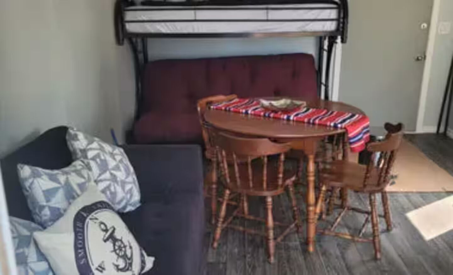 Une pièce avec un lit superposé, une table ronde en bois entourée de chaises, un canapé et un plaid à motifs posé sur la table.