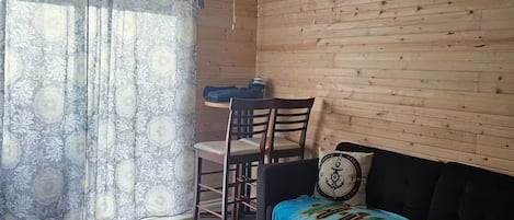 A room with a ceiling fan, a wooden wall, a patterned curtain, a dining table with chairs, and a sofa with a decorative pillow.
