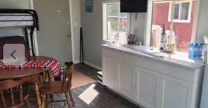 Ein Zimmer mit einem Etagenbett, einem Esstisch mit Stühlen, einer Küchenzeile mit Fenster und einem an der Wand montierten Fernseher.