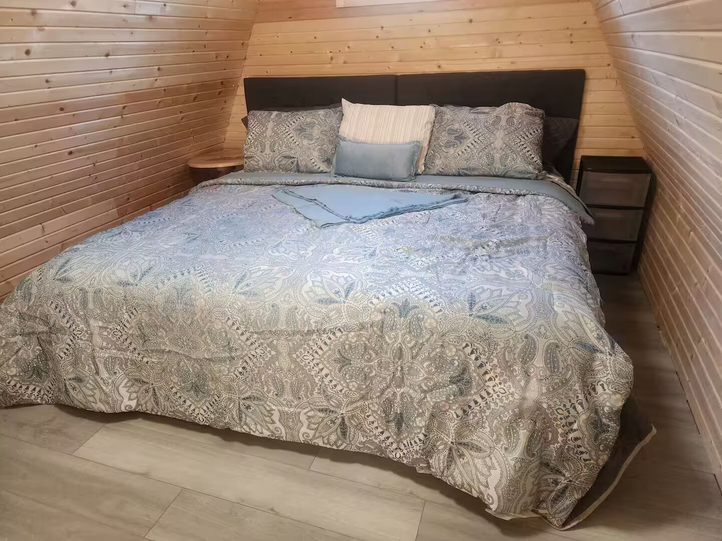 Quarto com teto inclinado de madeira, cama com roupa de cama estampada, mesa de cabeceira e uma pequena cômoda.