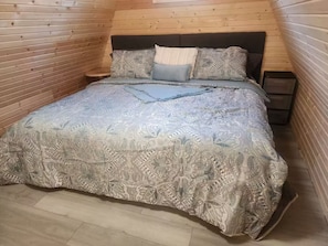 A bedroom with a wooden sloped ceiling, a bed with patterned bedding, a nightstand, and a small drawer unit.