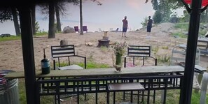An outdoor seating area with a table and chairs, a potted plant, and a view of a beach with people and chairs in the distance.