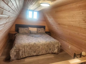 Un dormitorio con techo inclinado, paredes de madera, una cama con ropa de cama estampada, dos mesitas de noche y una ventana.