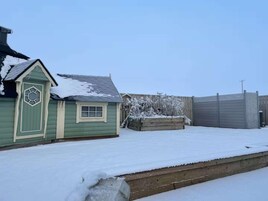 Un patio nevado con un cobertizo pequeño, una cerca de madera y un jardín elevado.
