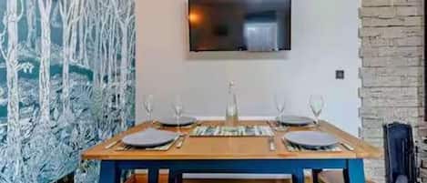 A dining area with a wooden table set for four, a flat-screen TV mounted on the wall, and a stone wall to the right.