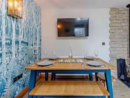Comedor con una mesa de madera dispuesta para cuatro personas, un televisor de pantalla plana en la pared y una pared de piedra a la derecha.
