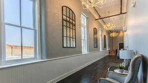 A hotel corridor with large windows, a chandelier, and a seating area with a lamp and plant. - The Royal on Main  (Batesville)
