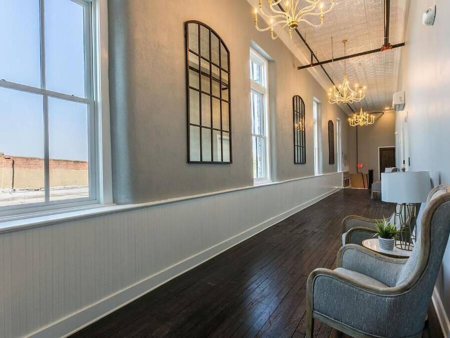A hotel corridor with large windows, a chandelier, and a seating area with a lamp and plant.