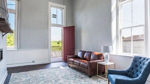 A room with a brown leather sofa, a blue armchair, a patterned rug, and large windows. - The Royal on Main  (Batesville)