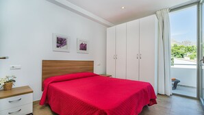 A bedroom with a bed, a nightstand, a dresser, and a window with a view of greenery. - Beachside Home With Shared Pool, Cala Molins (Pollença)