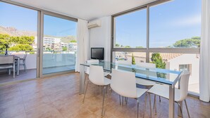 A modern living room with a glass dining table, white chairs, a flat-screen TV, and large windows offering a city view. - Beachside Home With Shared Pool, Cala Molins (Pollença)