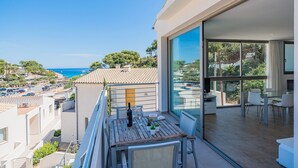 A balcony with a wooden table, a bottle, and glasses, overlooking a coastal town with buildings and a clear blue sky. - Beachside Home With Shared Pool, Cala Molins (Pollença)