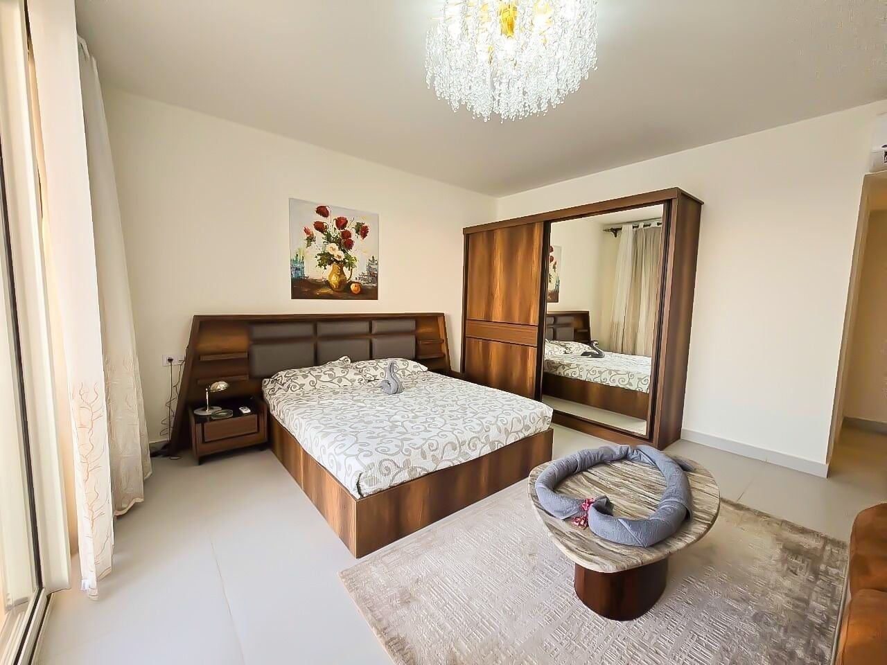 A bedroom with a large bed, a wardrobe, a mirror, a bedside table, and a decorative chair.