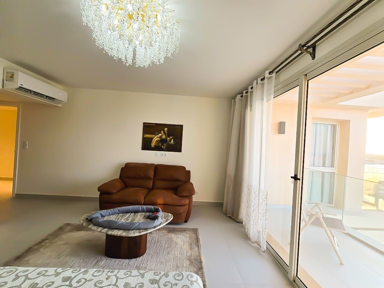 A living room with a brown sofa, a patterned rug, a small round table, a chandelier, and a sliding glass door leading to a balcony.