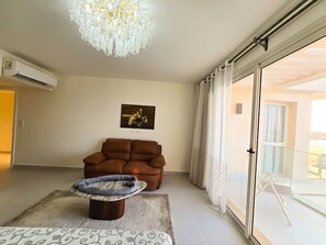 Ein Wohnzimmer mit braunem Sofa, einem gemusterten Teppich, einem kleinen runden Tisch, einer Chandelüster und einer Glastür zum Balkon.