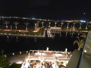 A nighttime view of a waterfront area with a dining area overlooking the water, palm trees, and distant city lights.