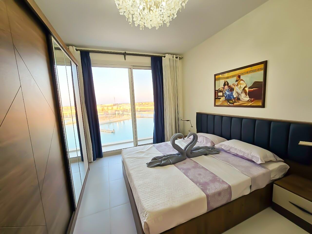 Ein Hotelzimmer mit einem großen Bett, einem Gemälde an der Wand und Blick auf ein Gewässer durch das Fenster.