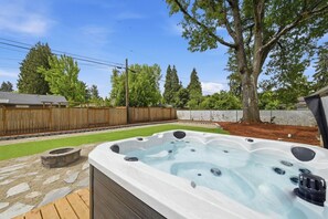 Un jacuzzi en un patio trasero con terraza de madera, un pozo de fuego de piedra y un área cercada con árboles.