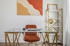 Ein moderner Arbeitsraum mit einem Glastisch, einem roten Sessel, einem Bücherregal mit goldener Rahmung und einem an der Wand hängenden abstrakten Gemälde.