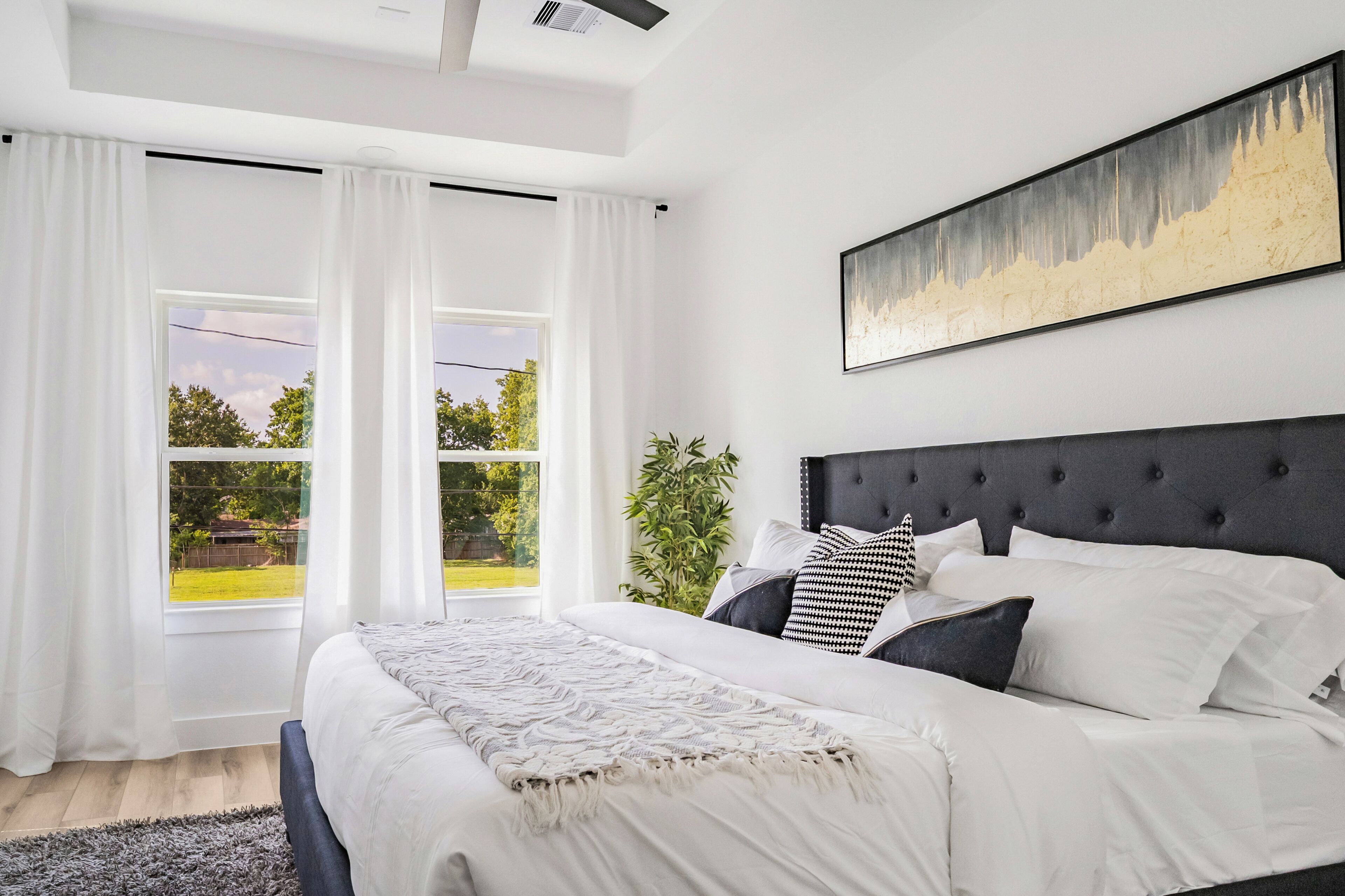 Un dormitorio con una cama grande, ropa de cama blanca, cabecera negra y una ventana con cortinas blancas.