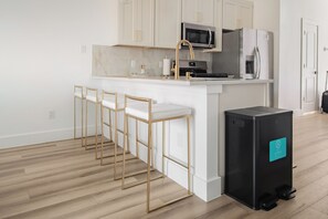 Een moderne keuken met witte kastjes, een roestvrijstalen koelkast, een magnetron en een koffieapparaat. Er staan drie barkrukken met gouden poten en witte kussens.